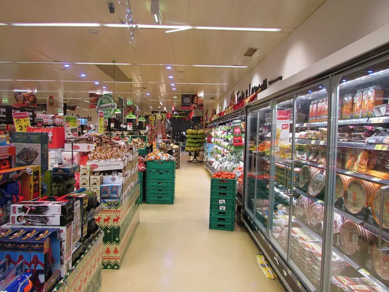 Corredor de um supermercado, com fruta à esquerda e frigoríficos à direita